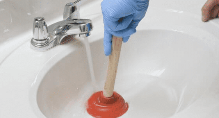 A person wearing a blue nitrile glove uses a red sink plunger to clear a drain in a white bathroom sink.