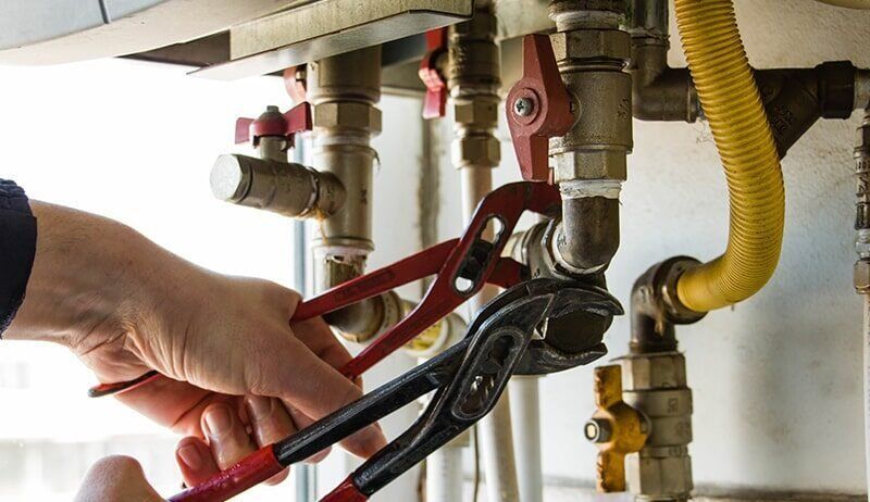 Close-up of hands using red channel-lock pliers to tighten a metal pipe fitting on a water heater system.
