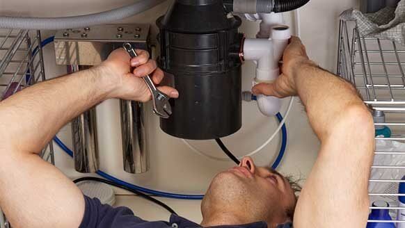 A person lying under a sink, using a wrench to repair a black garbage disposal unit.