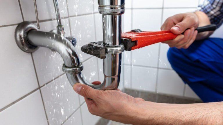 A plumber in blue work pants uses a red pipe wrench to tighten a chrome p-trap under a sink with white tiled walls.