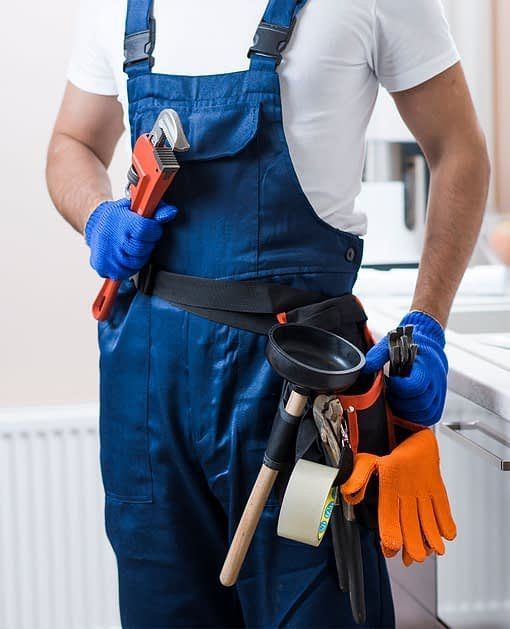 Plumber in blue overalls and gloves holding a pipe wrench, with tools and a plunger on a belt.