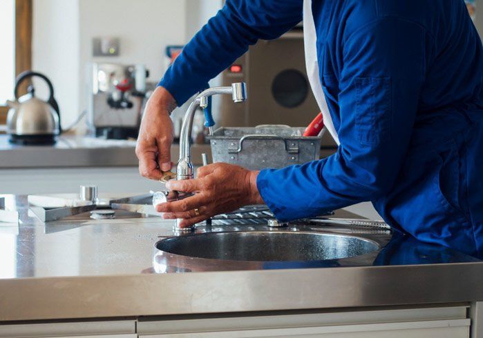 A person in a blue uniform repairing a kitchen faucet over a stainless steel sink.