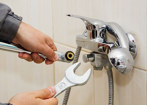 A person uses an adjustable wrench to repair a chrome shower faucet mounted on a tiled wall.