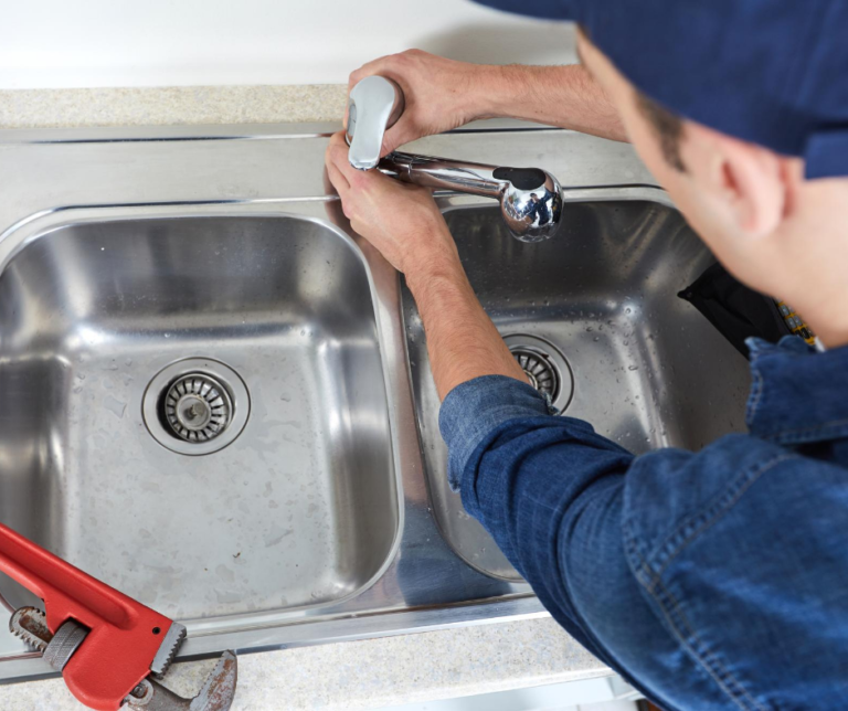 A plumber in a blue uniform repairing a stainless steel kitchen sink faucet, with a red pipe wrench nearby.