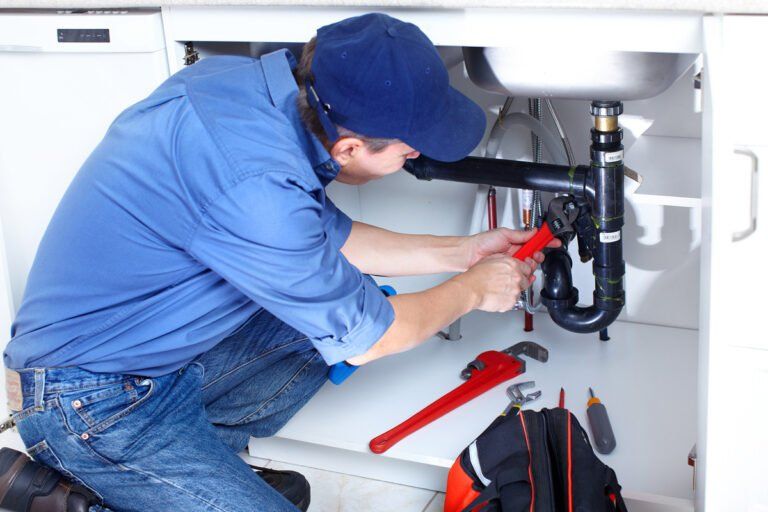 A plumber in a blue uniform kneels under a sink, using a wrench to repair the drain pipes.