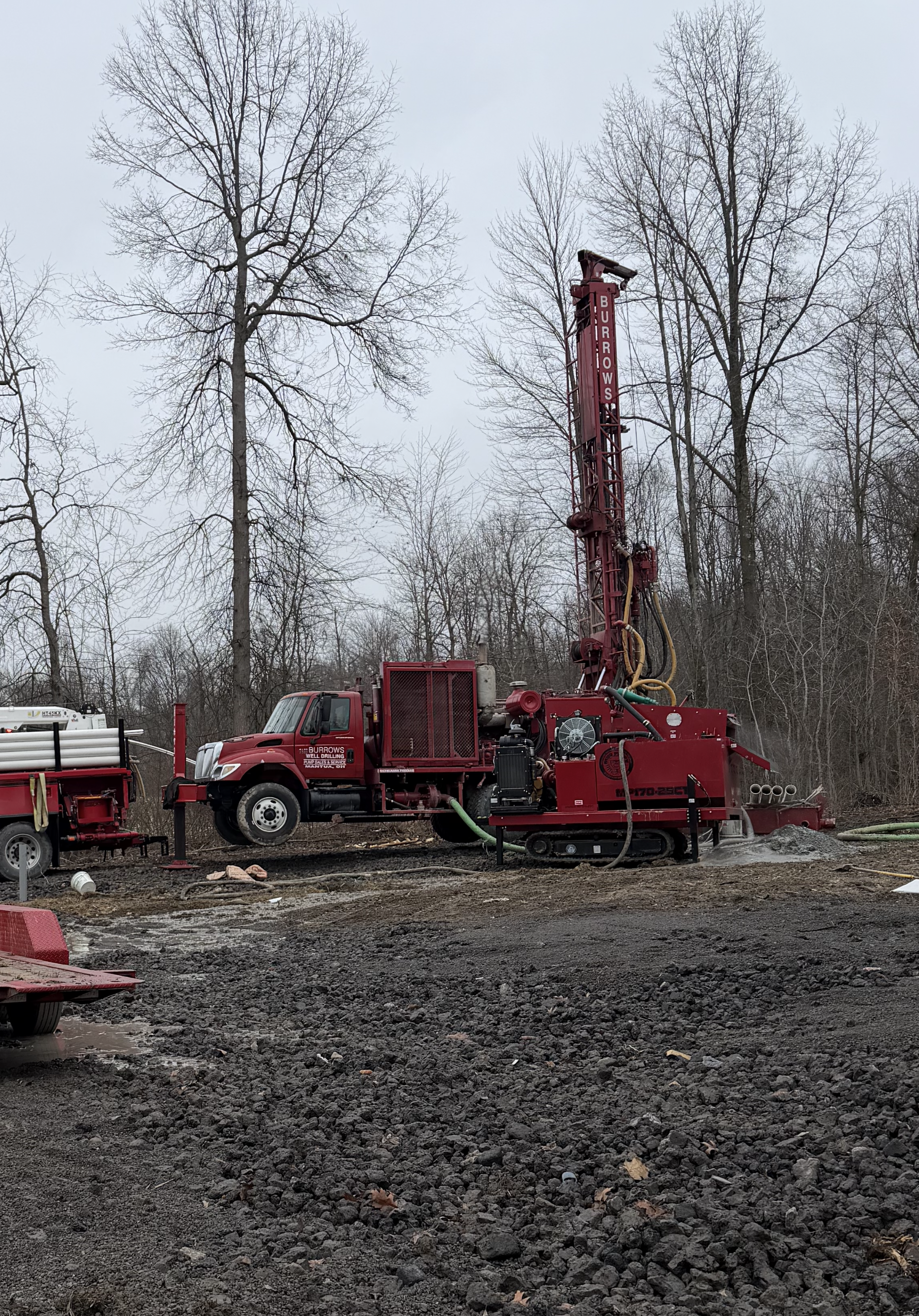 Red well drilling trucks in a grassy field; cloudy sky, trees in background.