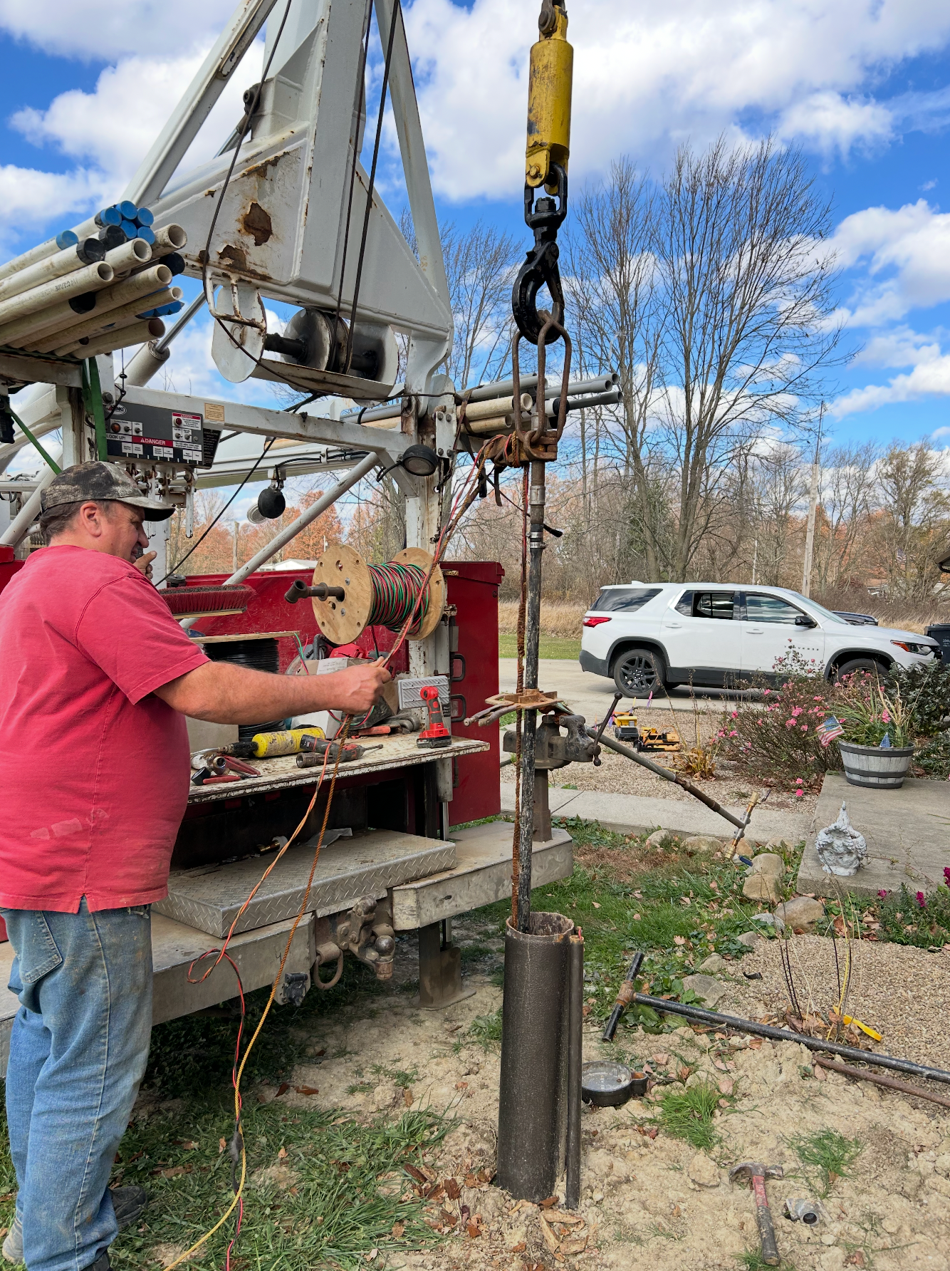 Man operating a drilling rig, preparing equipment. Outdoors, with a white truck and trees in the background.