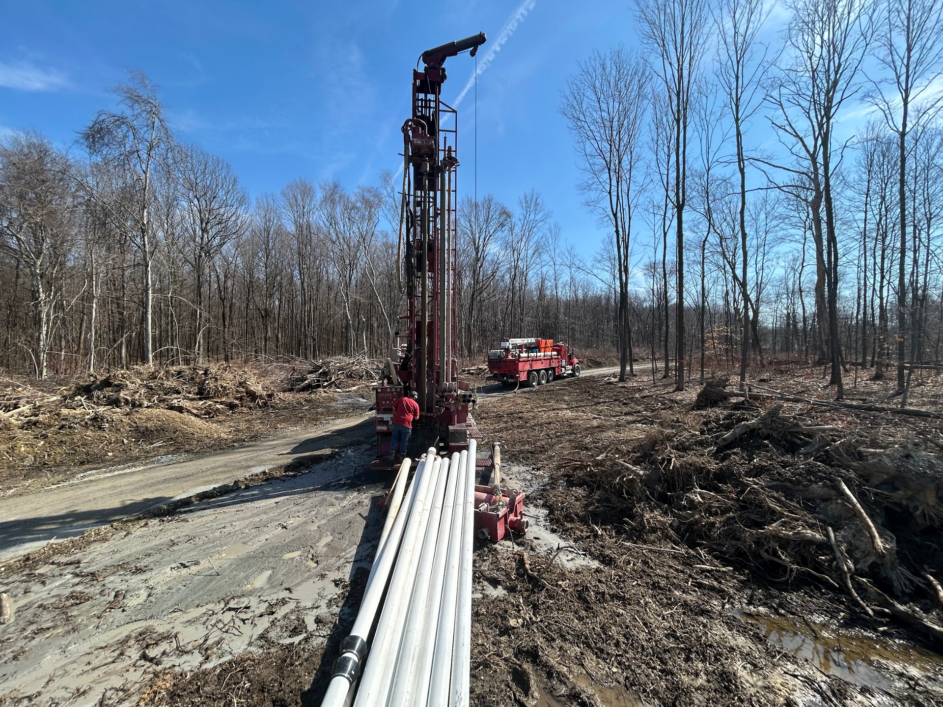 Drilling rig in a wooded area, with a truck in the background and a pile of white pipes in the foreground.