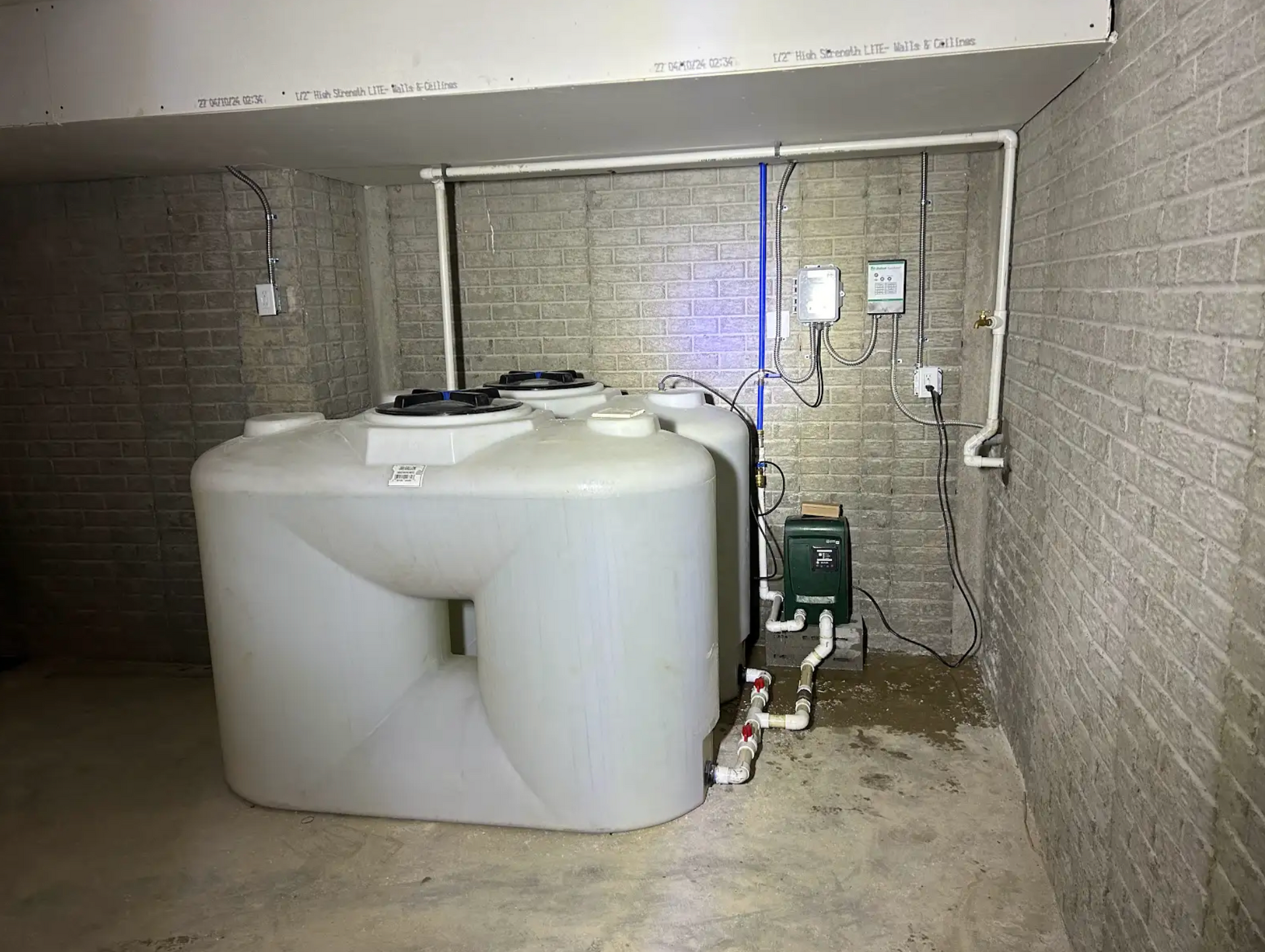 Large white water storage tank in a basement setting with associated pipes, controls, and brick walls.