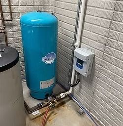 Blue well water storage tank with a pressure switch and piping, next to a white brick wall.