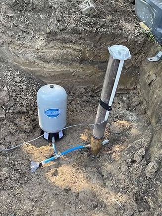 Well water system components in a dirt pit: well casing, pressure tank, and pipes.