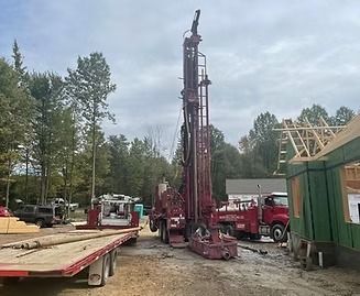 A drilling rig at a construction site with a flatbed trailer and a red truck.