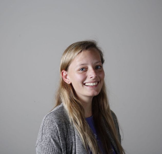 A woman is smiling for the camera while wearing a gray sweater and a purple shirt.