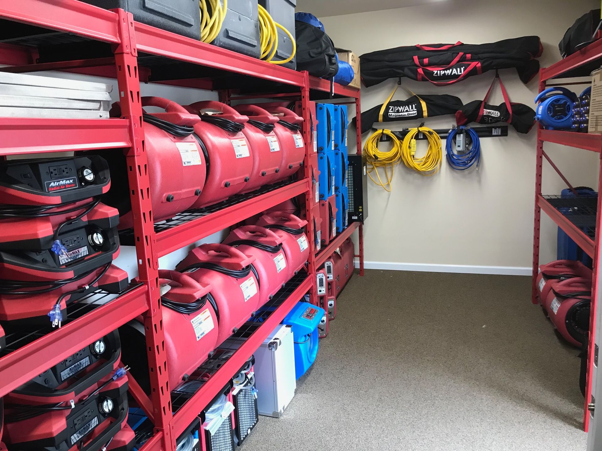 Storage shelves stocked with red industrial air movers, yellow power cords, and equipment bags in a room with carpet.