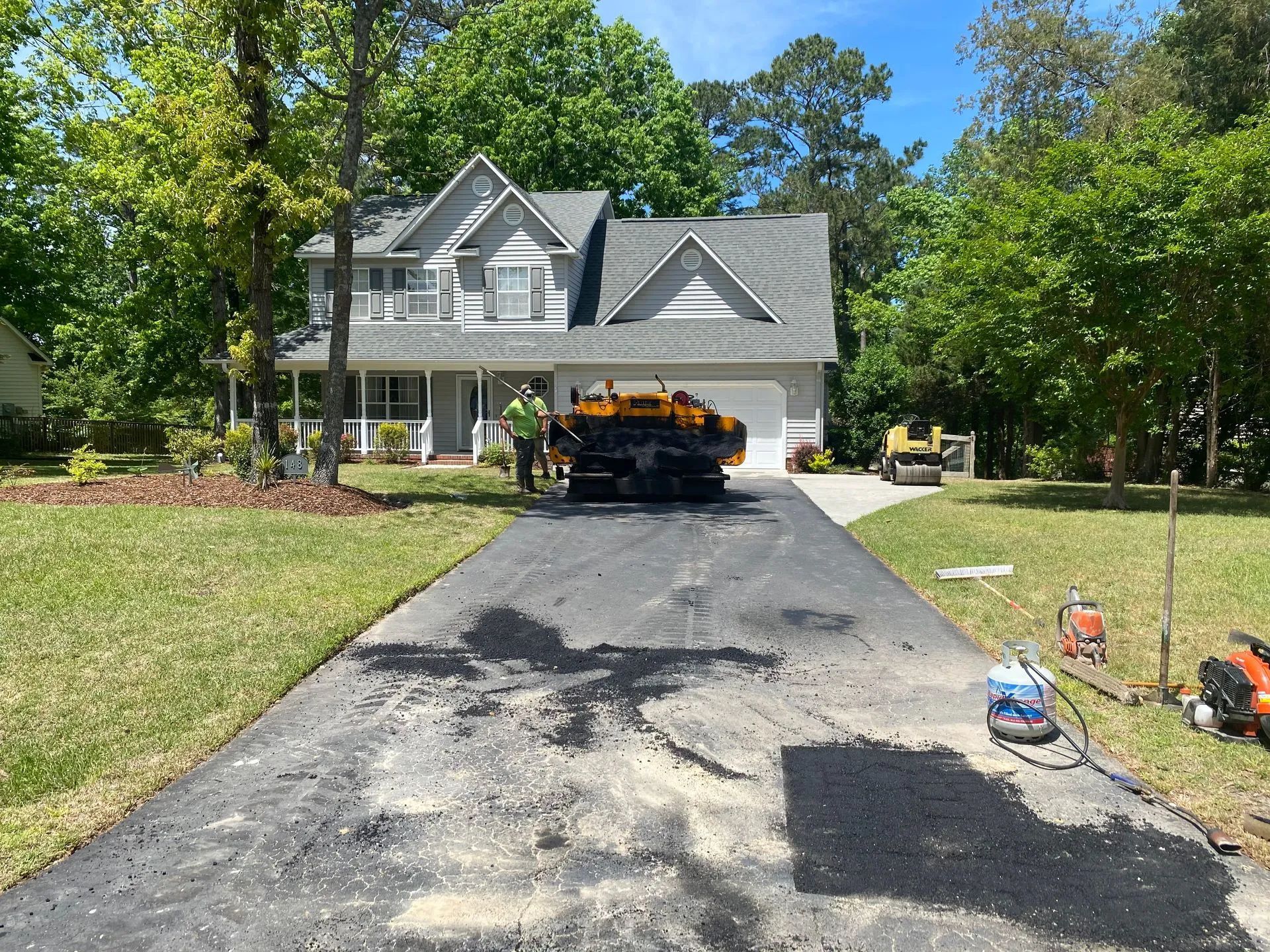 Asphalt driveway being paved at a house. Workers, machinery, and trees are visible.