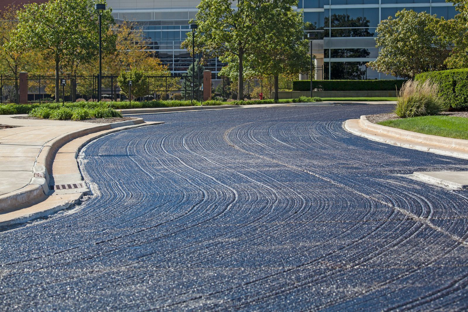 Freshly paved asphalt road curves near a building, trees, and landscaping.