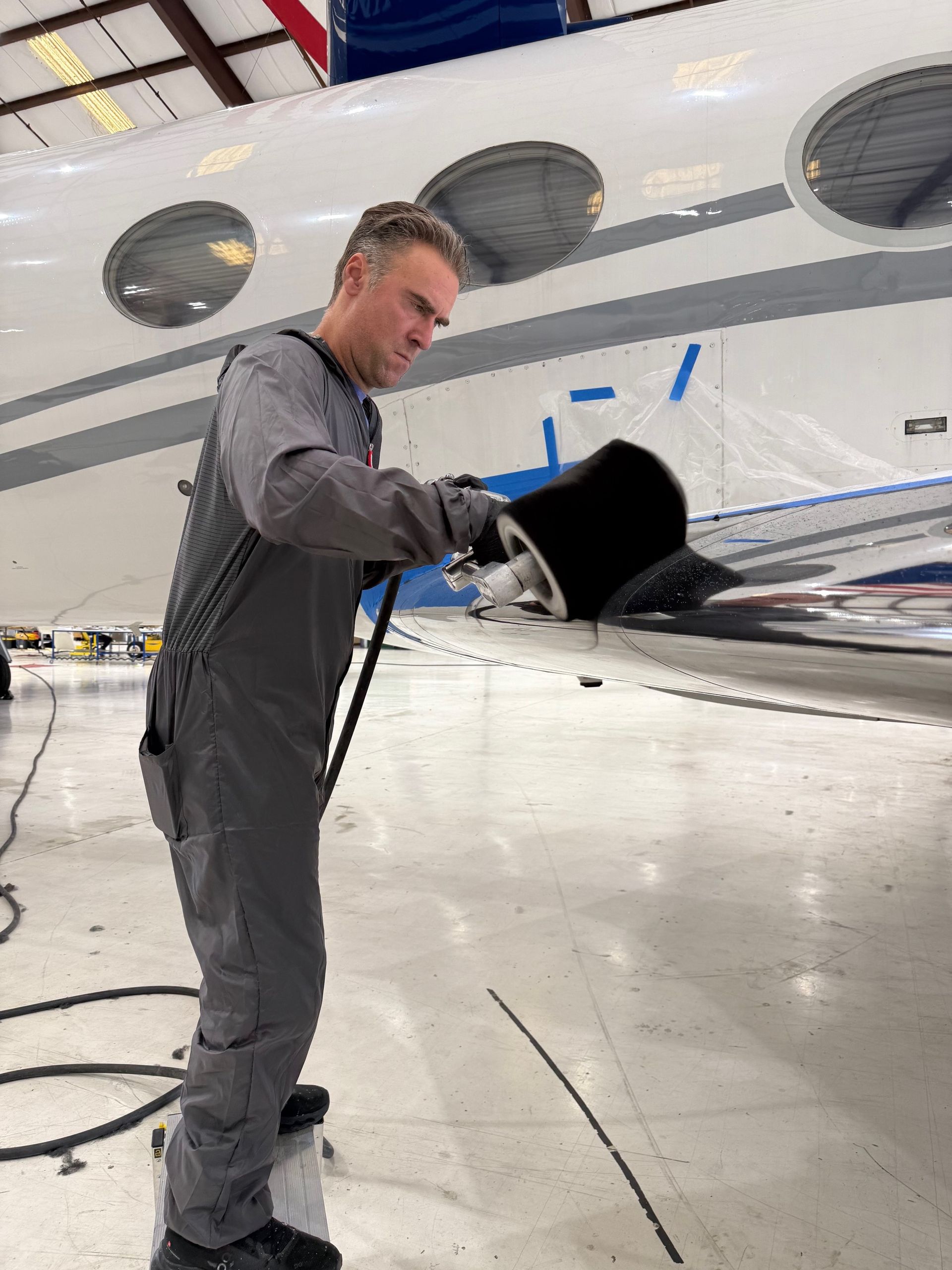 Mechanic in grey jumpsuit working on a plane wing, likely inspecting or cleaning in a hangar.