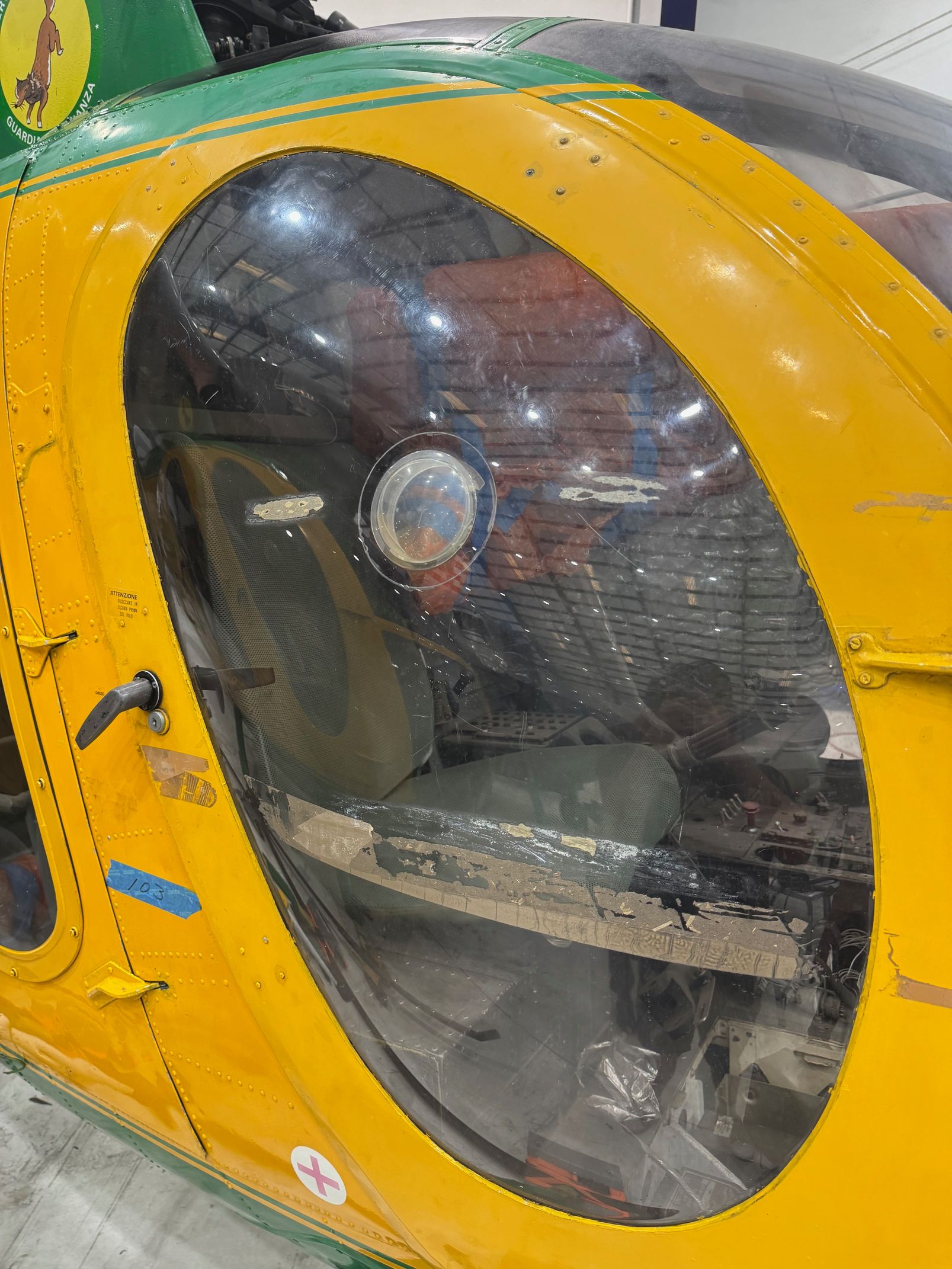 Yellow and green helicopter cockpit with a large, oval window. Interior visible through clear glass.