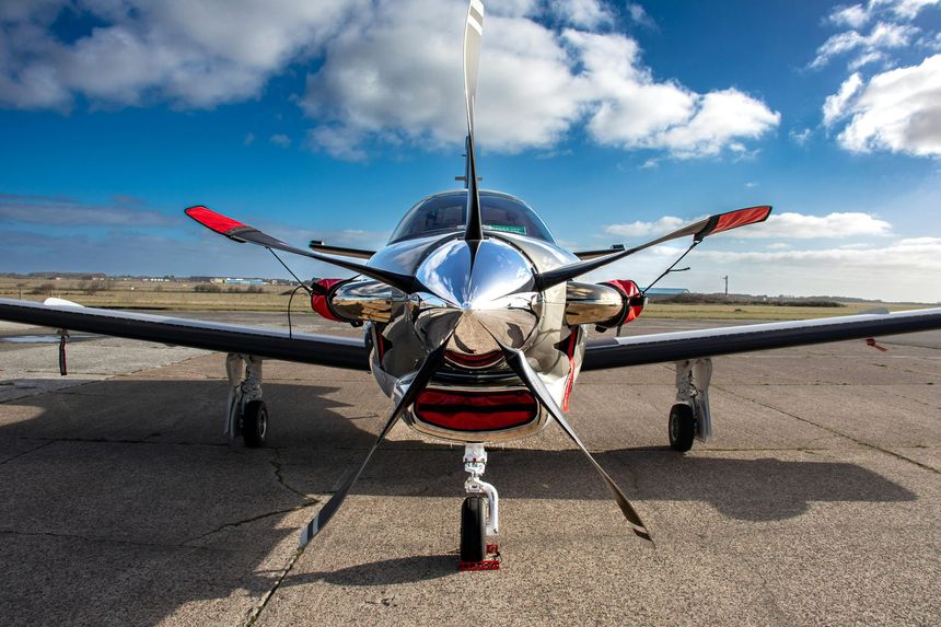 Silver airplane on tarmac, front view. Propeller blades and wing tips have red accents, against a blue sky with clouds.