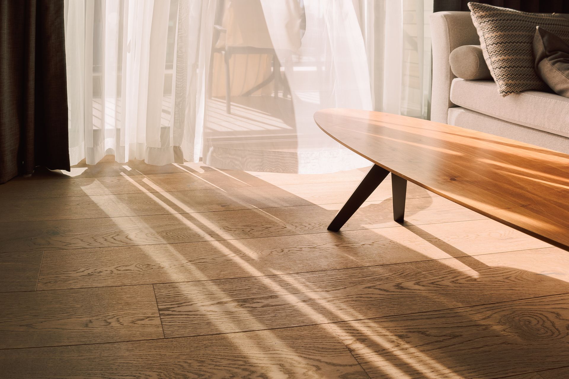 Sunlight streams through sheer curtains onto patterned flooring in a living room, highlighting a wooden coffee table.
