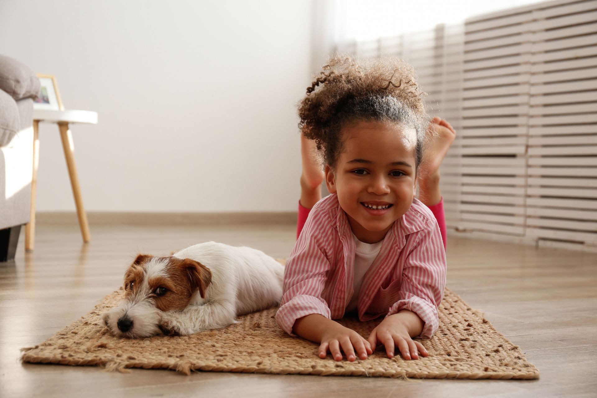 Girl smiling, lying on floor, with a dog resting beside her on a woven rug.