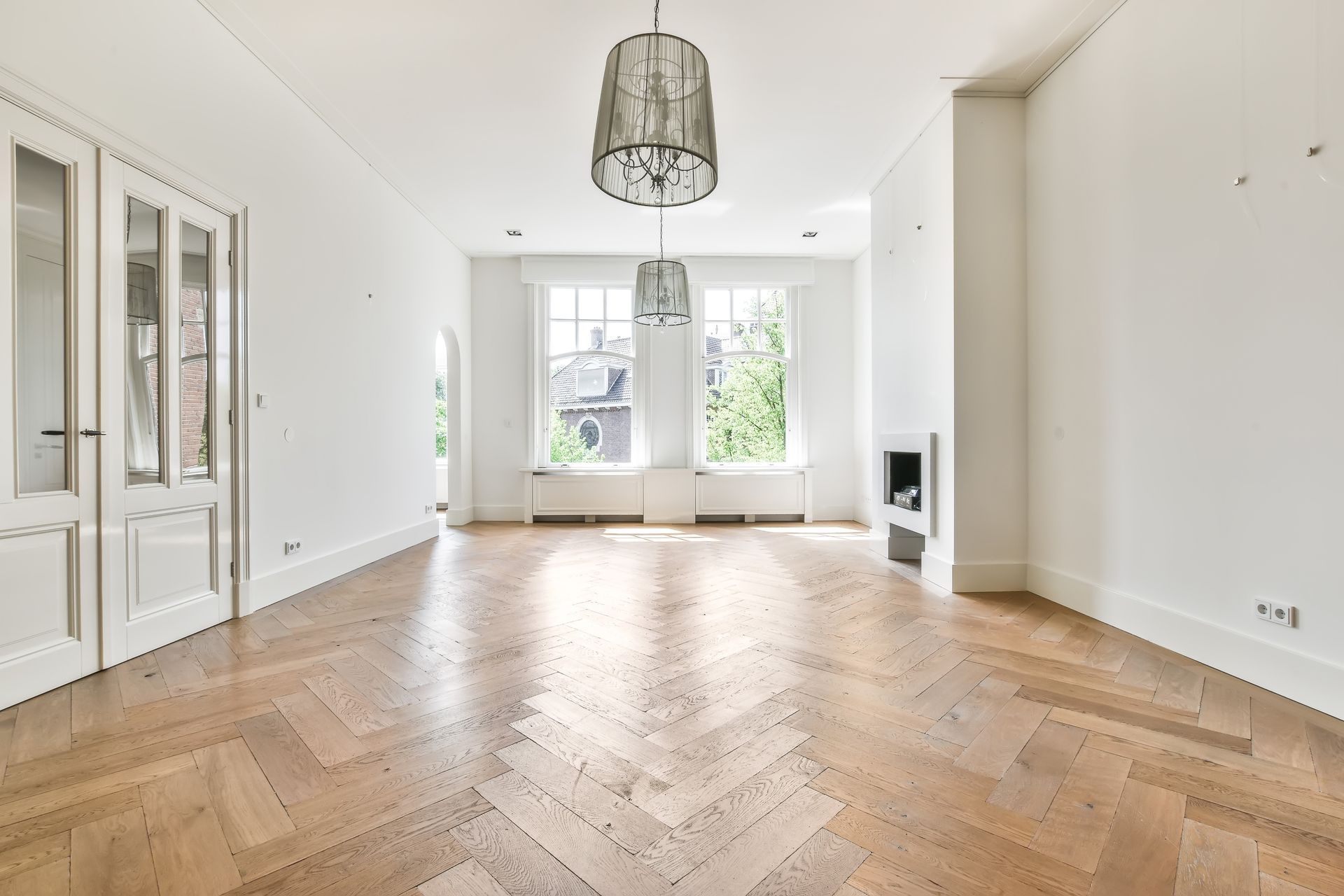 Empty room with herringbone wood floor, white walls, two windows, and hanging lights.