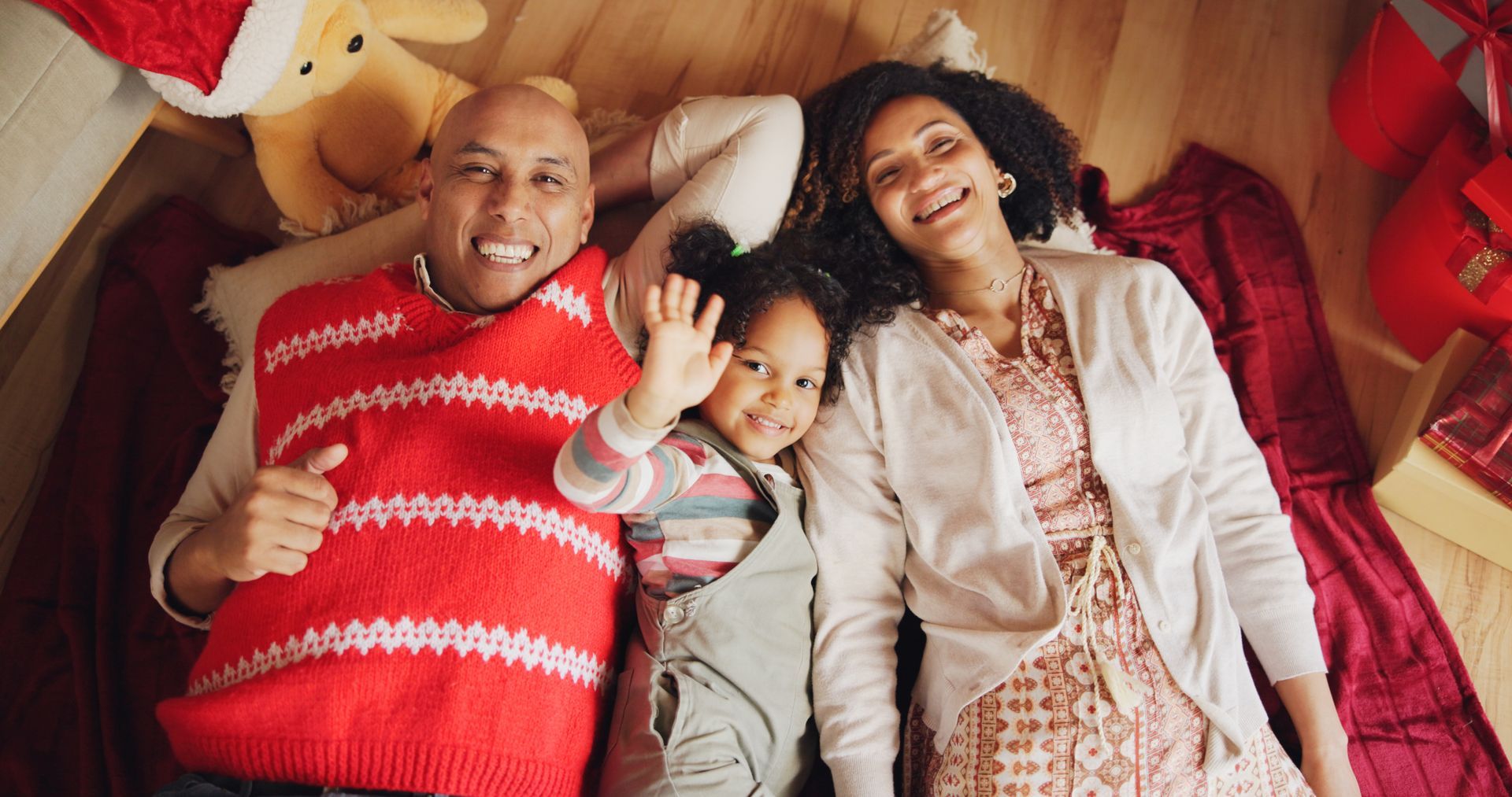 Family lies on floor, surrounded by gifts and decorations. The father gives a thumbs up, daughter waves, and mother smiles.