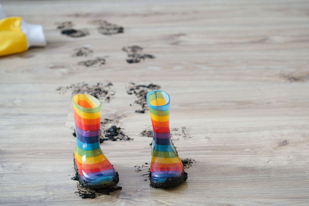 Rainbow-striped rubber boots stand on a wooden floor, surrounded by muddy footprints leading away from them.