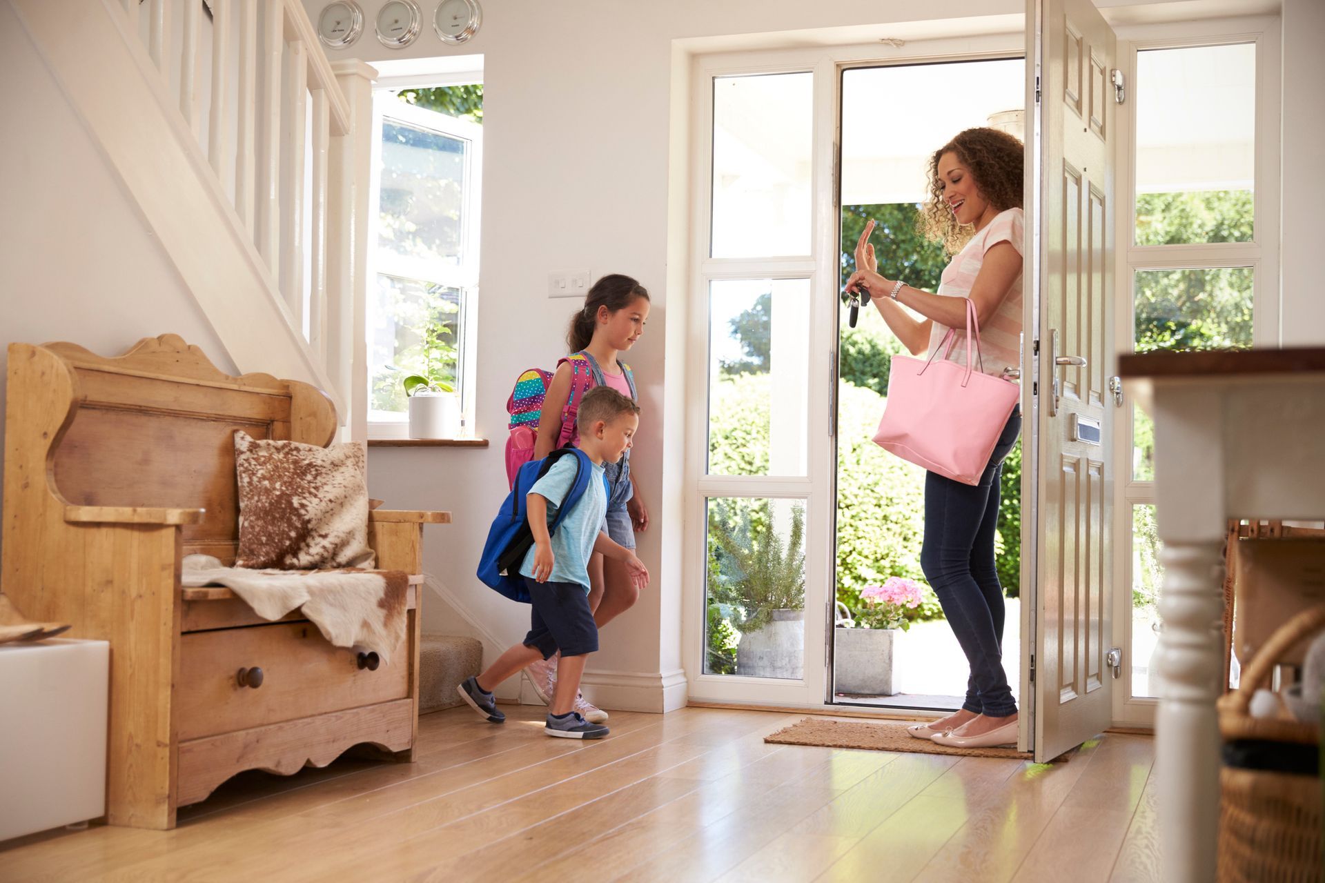 A woman waves goodbye to two children with backpacks as they exit the house. The scene is a bright, inviting entryway.