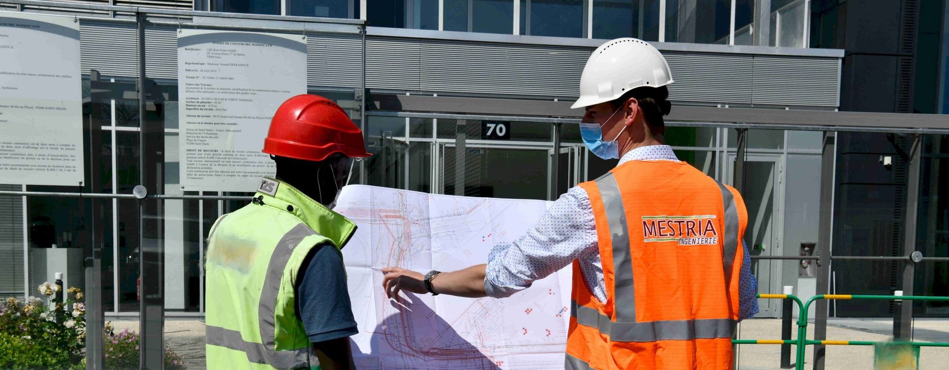 Deux ouvriers du bâtiment regardent un plan devant un bâtiment.