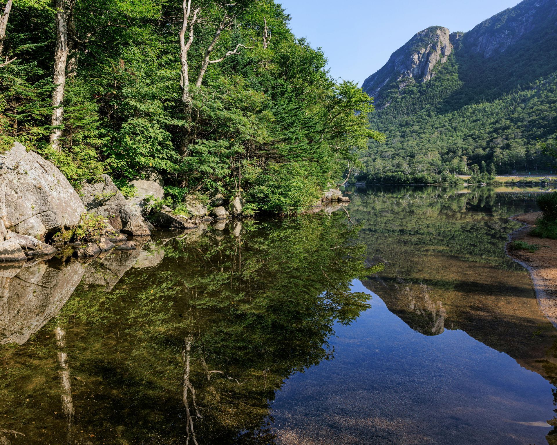 Calm lake reflects green trees and a mountain under a bright blue sky.