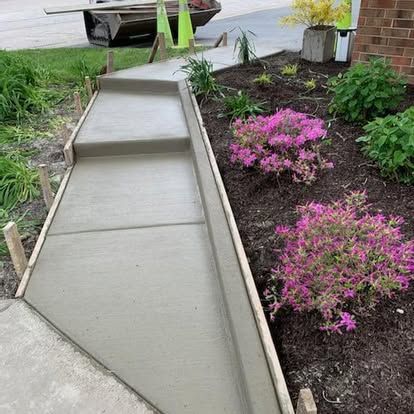 Freshly poured concrete walkway with wooden forms, next to a garden of pink flowering shrubs.