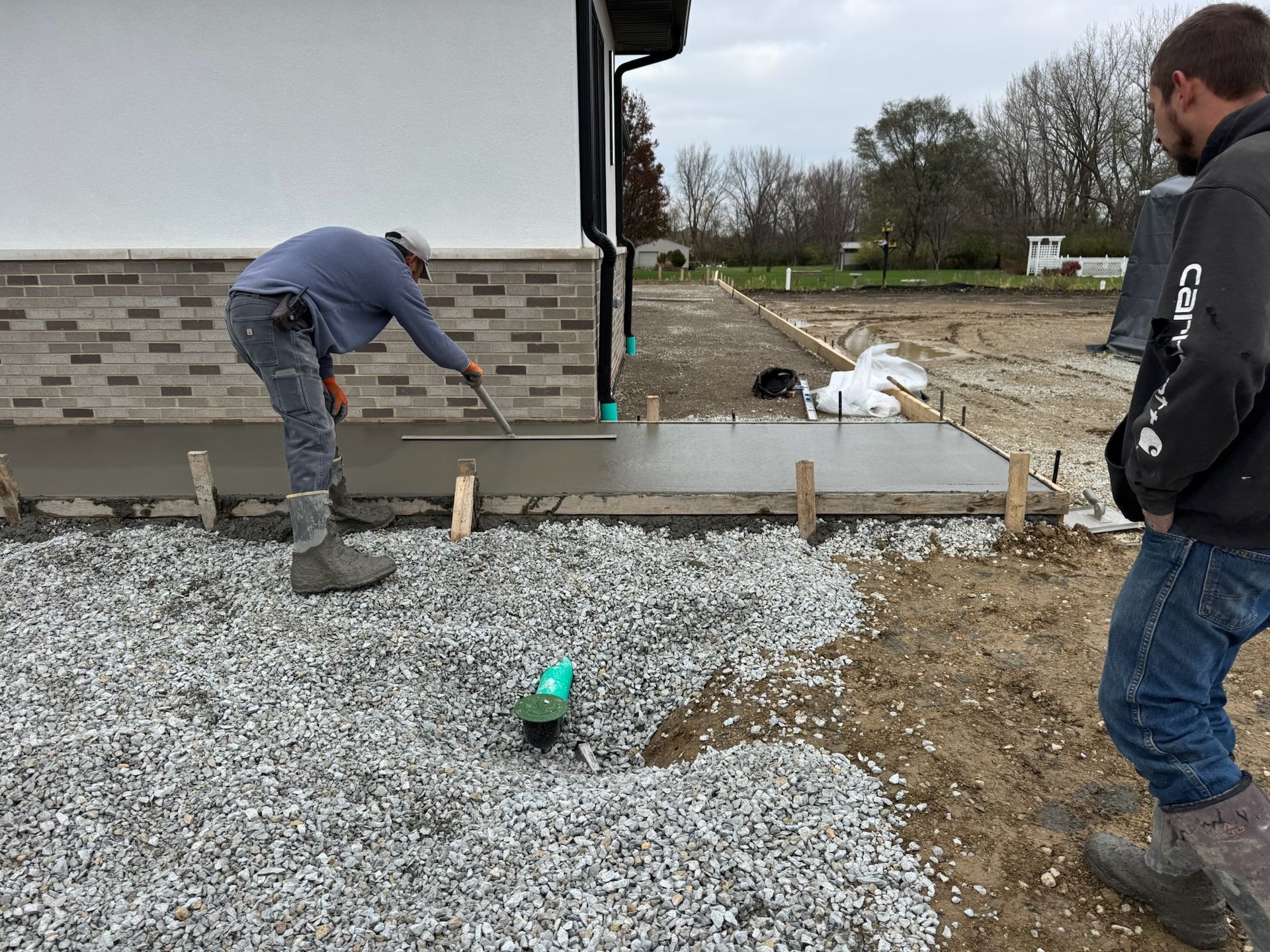 Two workers smoothing fresh concrete sidewalk. One uses a trowel. Gravel and dirt surround the work area.