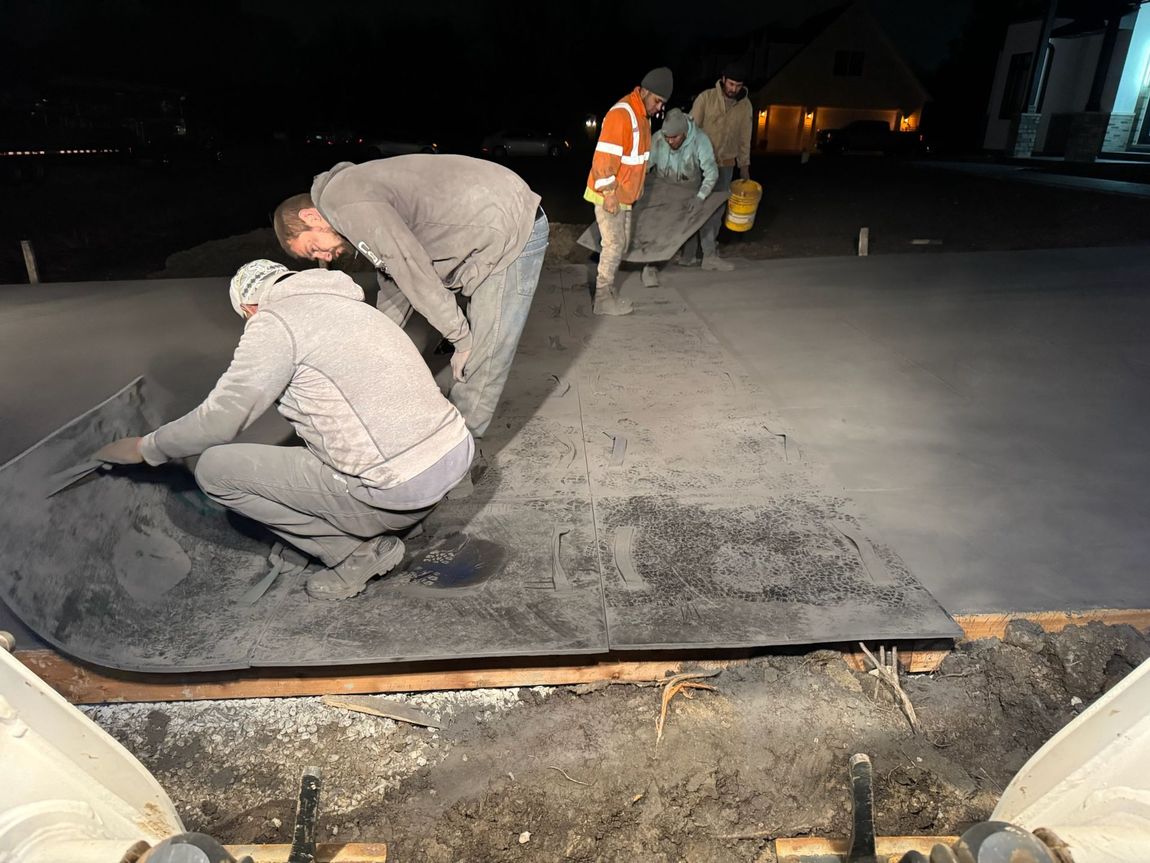 Workers smoothing concrete at night. One kneels, others carry a sheet and bucket in a dimly lit outdoor setting. Workers smoothing concrete at night. One kneels, others carry a sheet and bucket in a dimly lit outdoor setting.