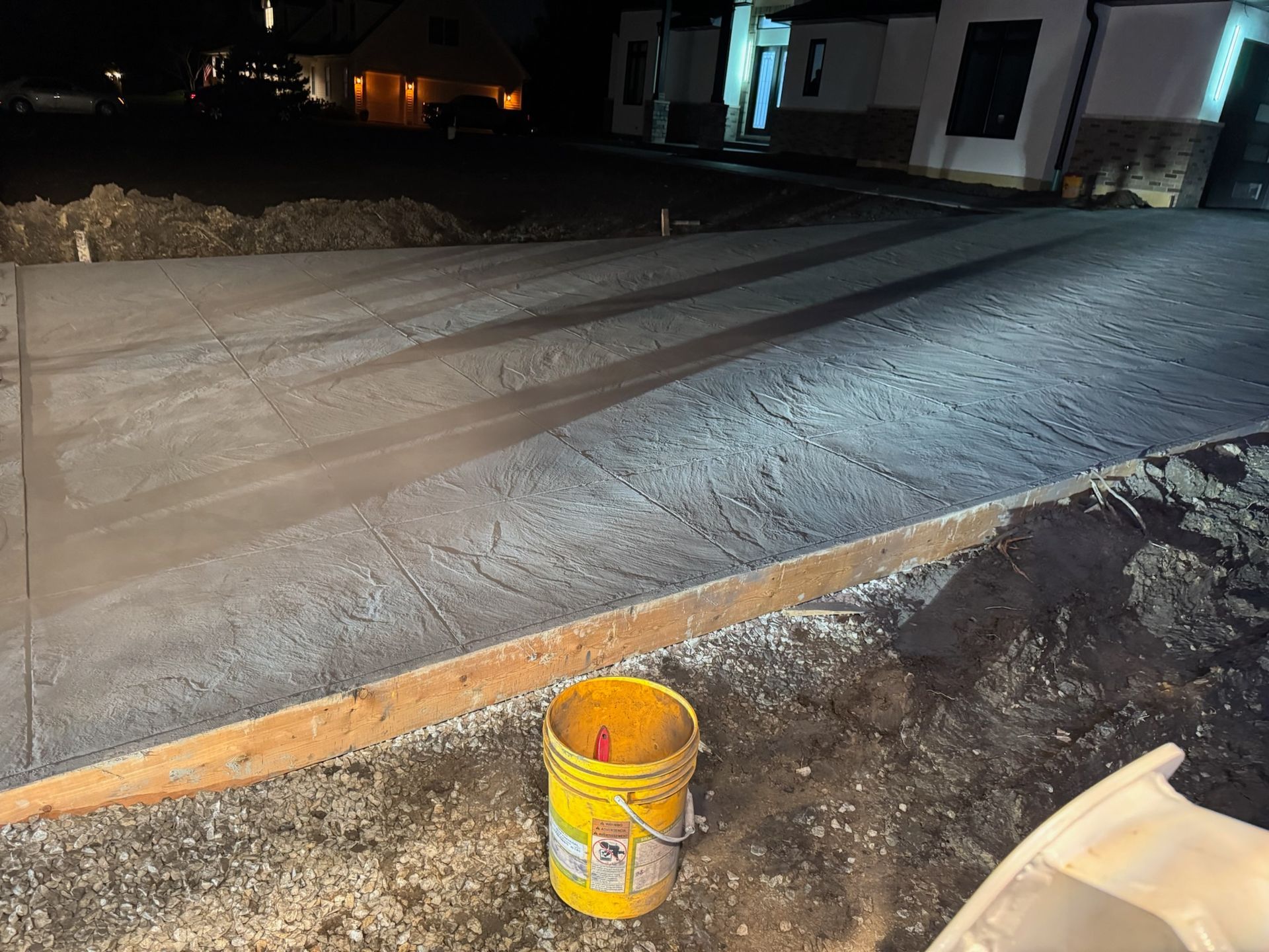Newly poured concrete driveway with wooden form, yellow bucket in foreground.