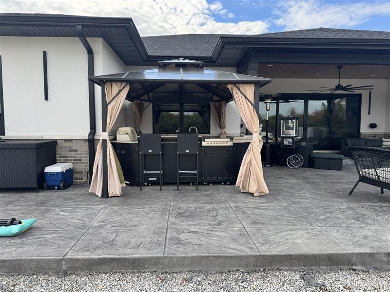Outdoor kitchen with a black frame and beige curtains on a gray patio.