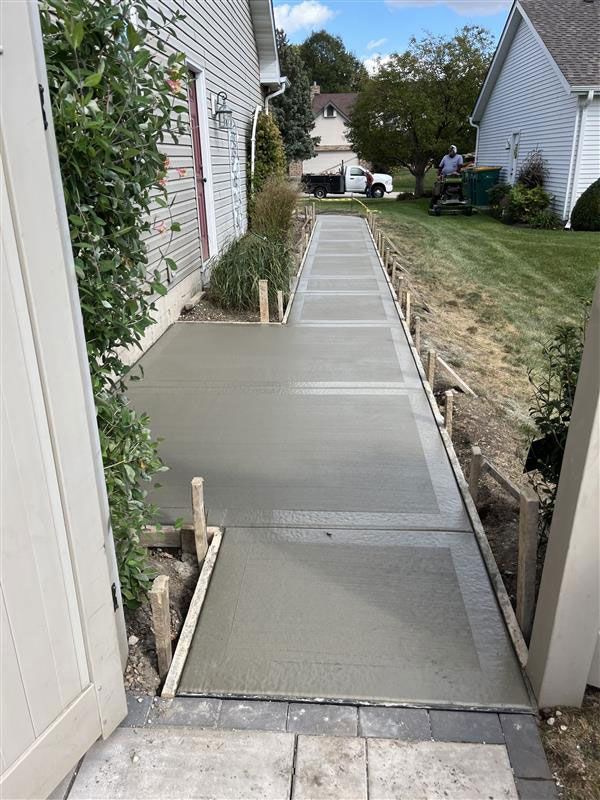 Newly poured concrete walkway next to a house with wooden formwork and landscaping.