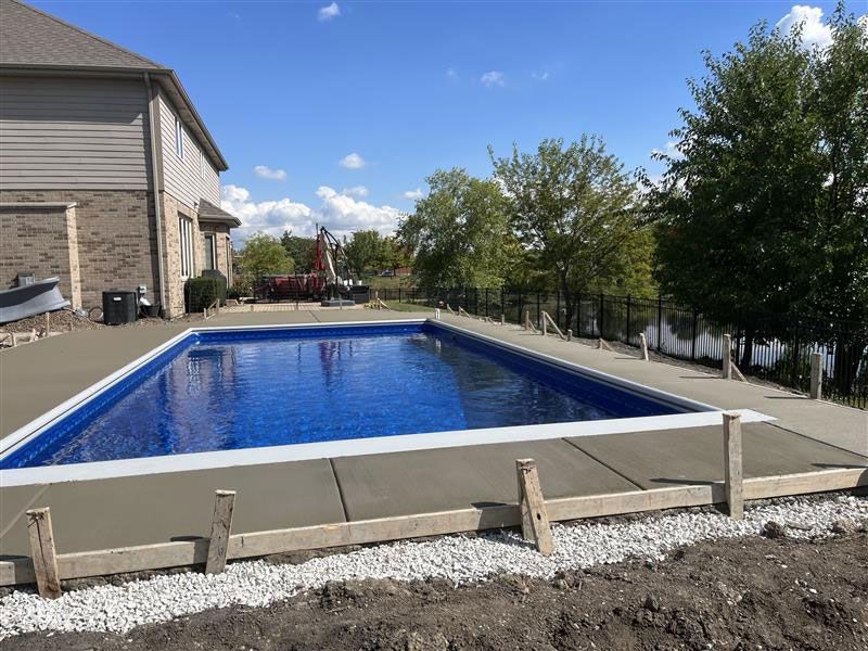 Rectangular swimming pool with concrete surround; a house is to the left, and trees are in the background.