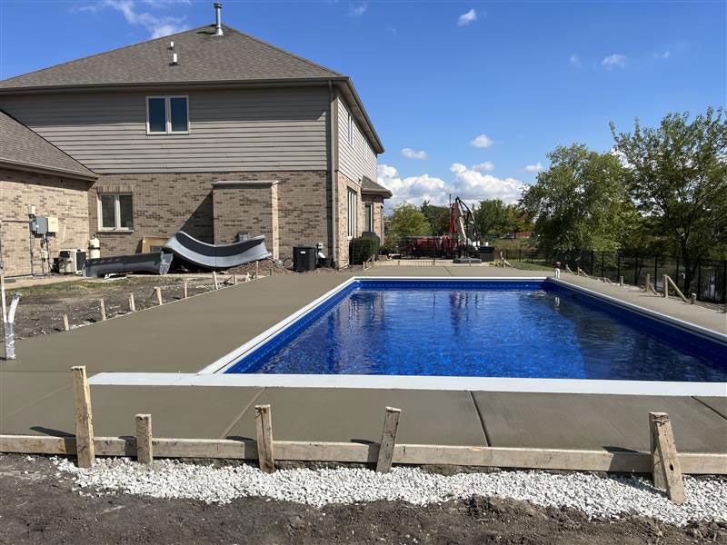 Pool with concrete decking being installed next to a two-story house, under a blue sky.