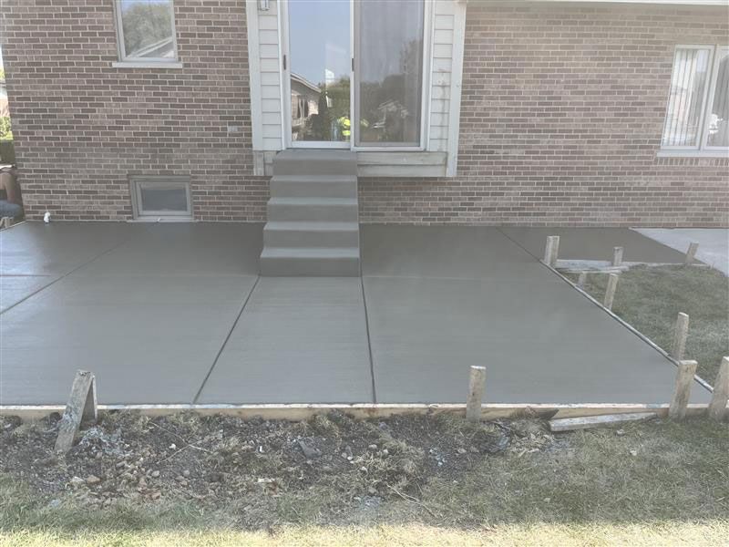 Newly poured concrete patio outside a brick building. Steps lead up to sliding glass doors.