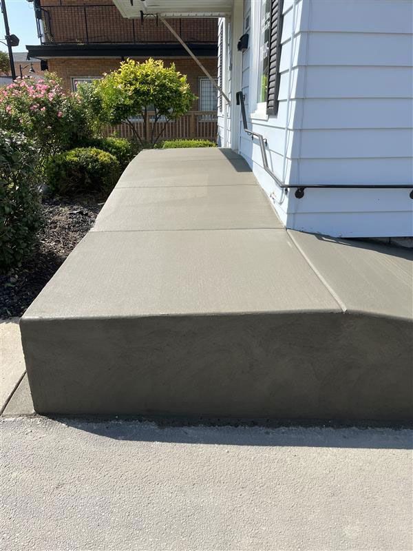 New concrete front porch and walkway next to a white house; gray concrete, green shrubs.