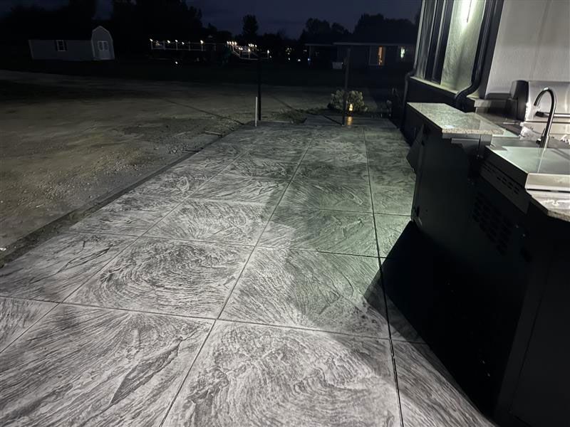 Exterior concrete patio with a stamped pattern, next to a dark outdoor kitchen, at dusk.