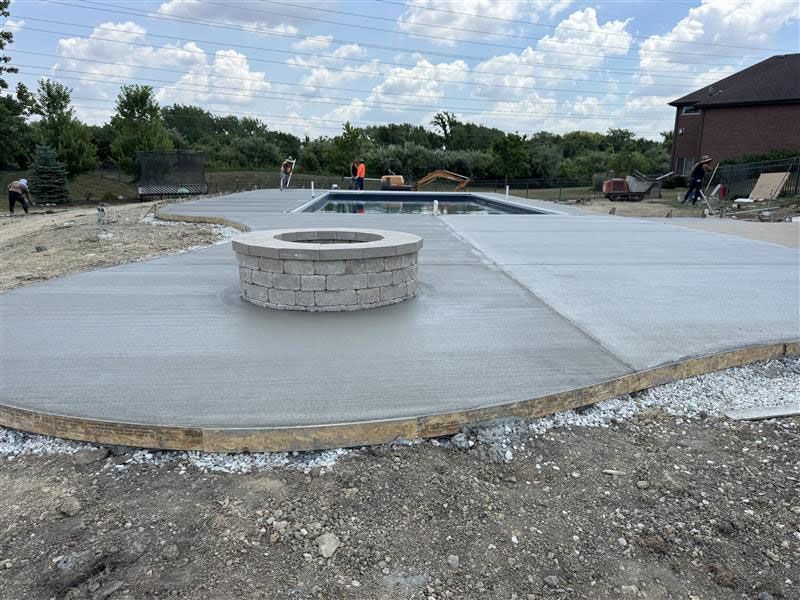 Newly poured concrete patio surrounding a swimming pool with a fire pit; construction in progress.