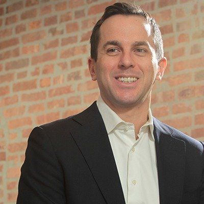 A man in a suit and white shirt is smiling in front of a brick wall