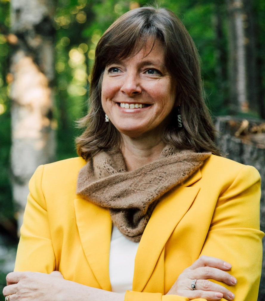 A woman in a yellow jacket and scarf is smiling with her arms crossed.