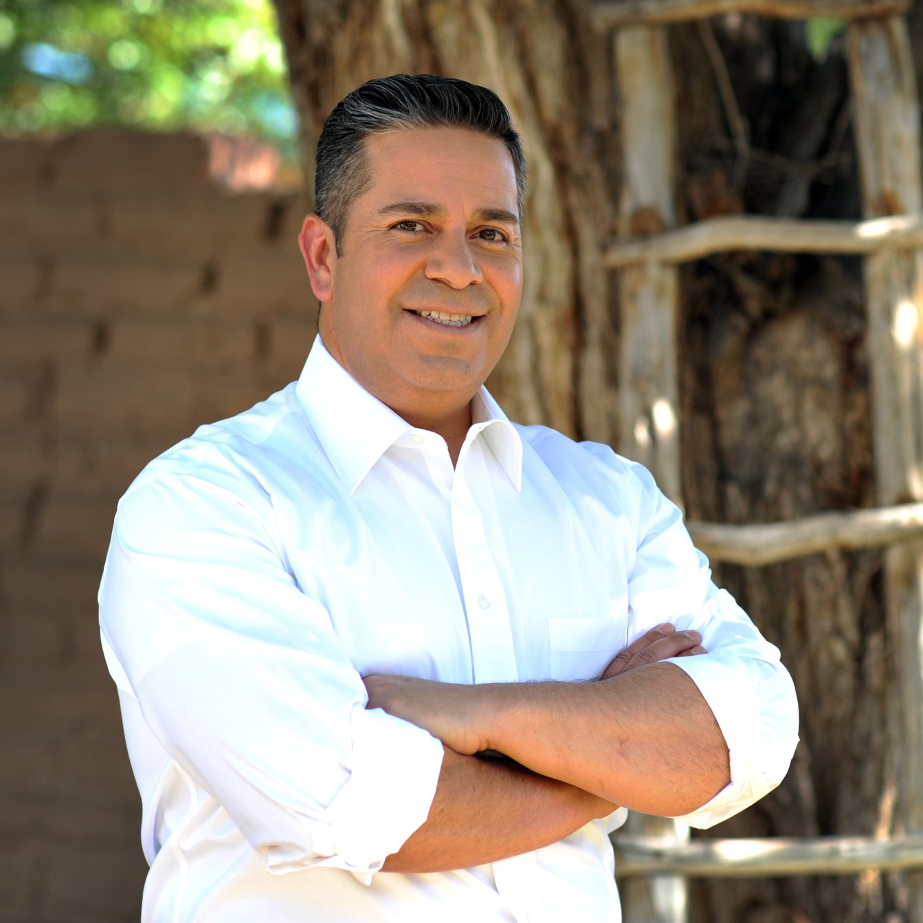 A man in a white shirt stands with his arms crossed in front of a tree