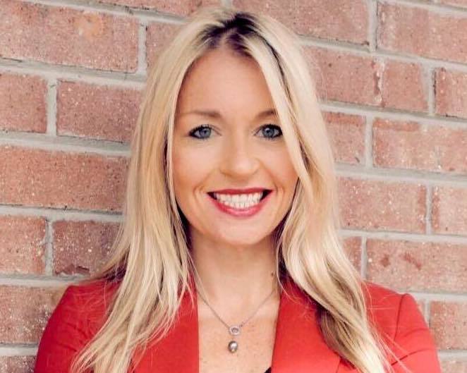 A woman in a red jacket is smiling in front of a brick wall.