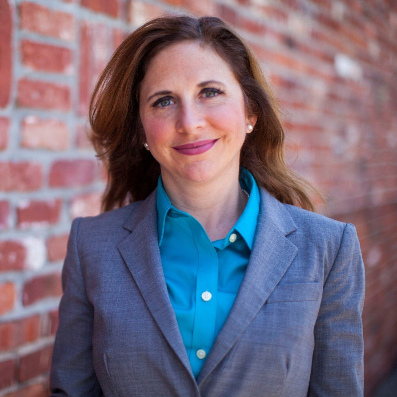 A woman in a suit and blue shirt is standing in front of a brick wall.