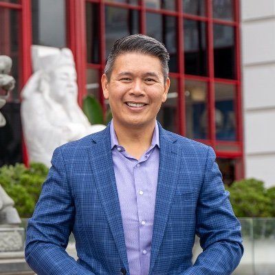 A man in a blue suit and purple shirt is standing in front of a building.