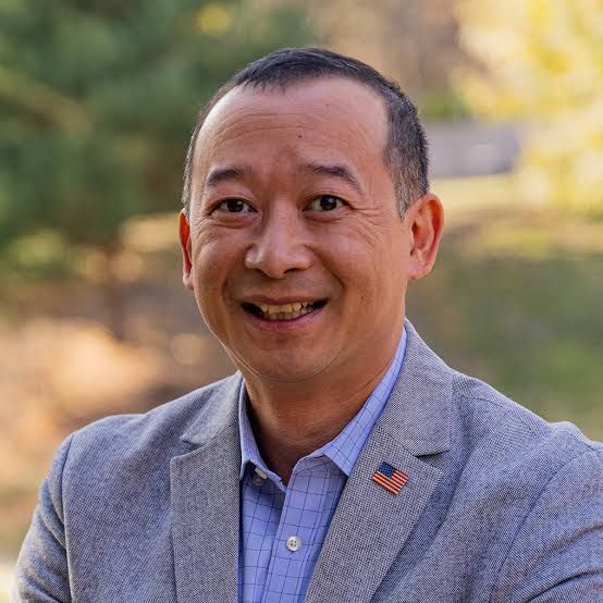 A man in a suit with an american flag pin on his jacket is smiling for the camera.